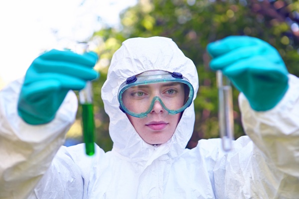 woman with blue goggles and gloves holding two graduated cylinders one on the left(clear liquid) and right(green liquid) hand.
