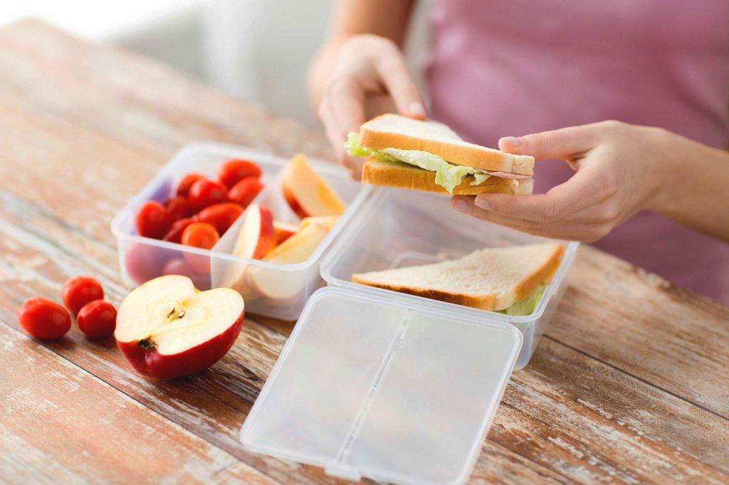 A woman standing in a kitchen holding a sandwich places it into a container next to an apple and strawberries.